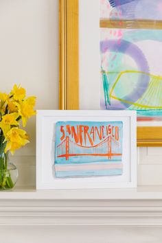 yellow flowers are in a vase next to a framed painting on a mantelpiece