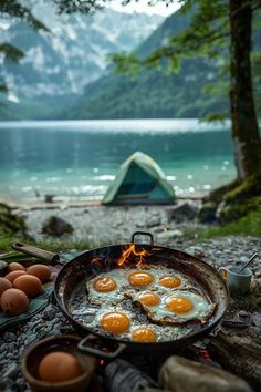 eggs are cooking on an outdoor grill in front of a lake and camping gear with a tent in the background