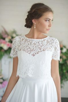 a woman wearing a white dress with lace on the top and sleeves, standing in front of flowers