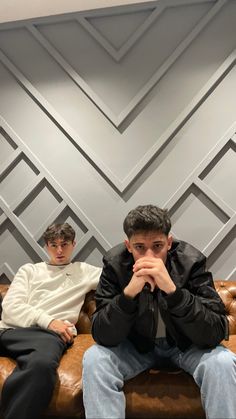 two young men sitting on a couch in front of a wall with geometric design behind them