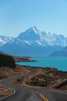 an empty road with mountains in the background and blue water on either side that is surrounded by brown grass