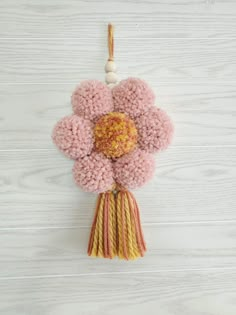 a pink and yellow flower hanging from a string on a white wooden wall with a tassel