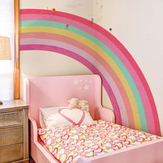 a child's bed with a rainbow painted on the wall behind it and a teddy bear