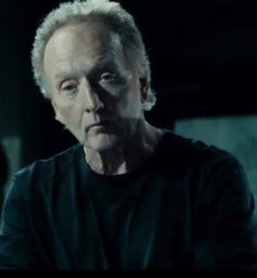 an older man with white hair and black shirt looks at the camera in a dark room