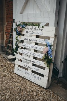 a wooden sign with flowers on it sitting in front of a white building and brick wall