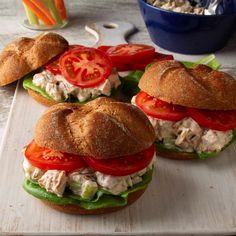 three chicken salad sandwiches with tomatoes and lettuce on a cutting board next to a bowl of carrots