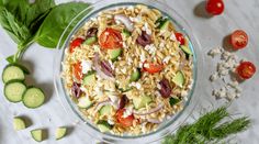 a glass bowl filled with pasta salad next to cucumbers, tomatoes and parsley