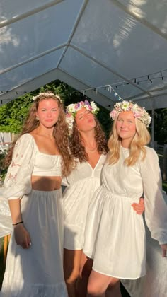 three women in white dresses are standing under a tent with flowers on their head and arms