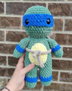 a hand holding a small crocheted turtle toy in front of a brick wall