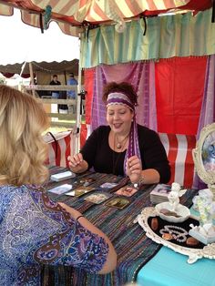 two women are sitting at a table with tea cups and plates on it, talking to each other