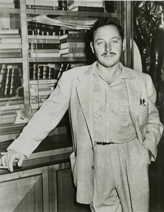 black and white photograph of man in suit with arms out, standing next to bookshelf