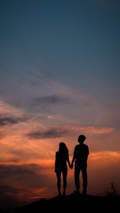 two people holding hands while standing on top of a hill with the sun setting in the background