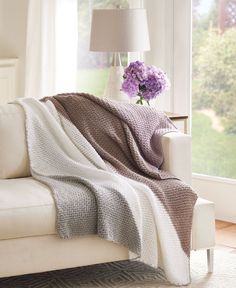 a white couch with a blanket on top of it next to a vase filled with purple flowers