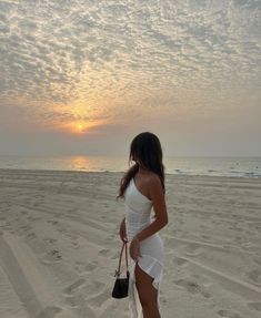 a woman standing on top of a sandy beach next to the ocean in front of a sunset