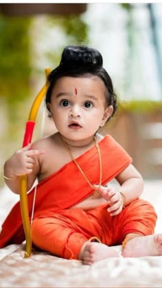 a baby girl dressed in an orange outfit holding a yellow stick and looking at the camera