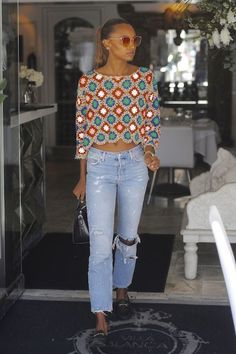 a woman wearing ripped jeans and a crop top is walking out of a building with her hand in her pocket