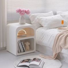 a bed with white sheets and pillows next to a book shelf on the floor in front of a window