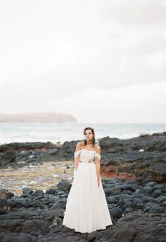 a woman in a white dress standing on rocks near the ocean with her arms behind her back