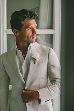 a man in a white suit and flower boutonniere looks out the window