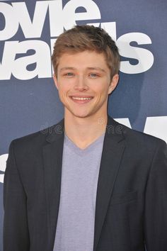 a smiling young man in a suit and t - shirt poses for the camera on the red carpet