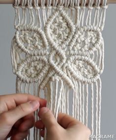 two hands are working on a macrame weaving project with white yarn and wood sticks