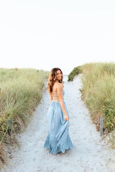 a woman in a blue dress is walking down a path by the beach with tall grass