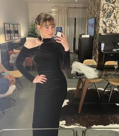 a woman in a black dress taking a selfie with her cell phone while standing in a living room