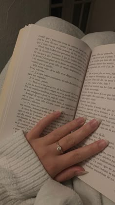 a woman's hand holding an open book in front of her face and wearing a ring