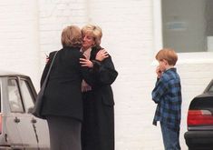 two women and a boy are hugging in front of a white brick building with cars