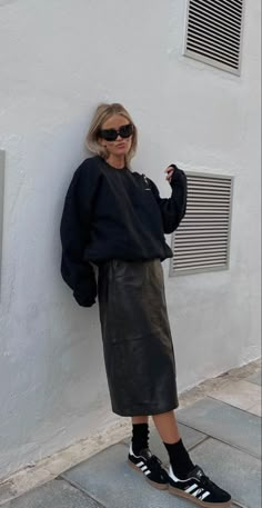 a woman leaning against a wall with her hand on her hip and wearing black sneakers