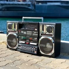 an old fashioned radio sitting on top of a brick sidewalk next to the water with a boat in the background