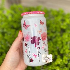 a hand holding a glass with pink flowers and butterflies painted on it, in front of some bushes