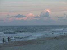 people are walking on the beach near the ocean at sunset or sunrise, with clouds in the sky