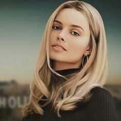 a woman with long blonde hair and big hoop earrings looking at the camera while standing in front of a cityscape