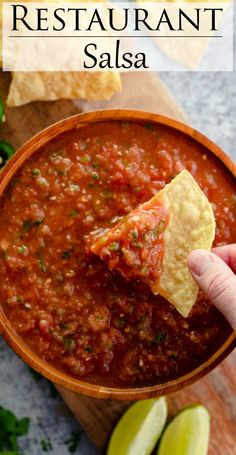 a hand dipping a tortilla chip into a bowl of salsa with limes and cilantro on the side