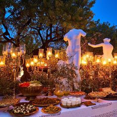 a table topped with lots of food and lit candles next to a statue in the background