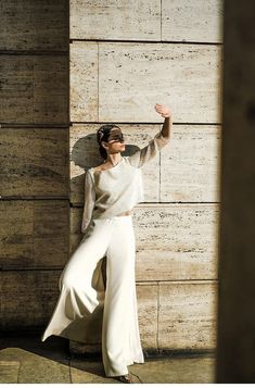 a woman leaning against a stone wall with her arms in the air and wearing white pants