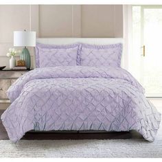 a bed with purple comforter and pillows in a room next to a lamp on a table