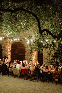 a group of people sitting at tables in front of trees with lights strung over them