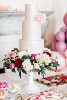 a wedding cake with pink and white flowers on it sitting on a table in front of balloons