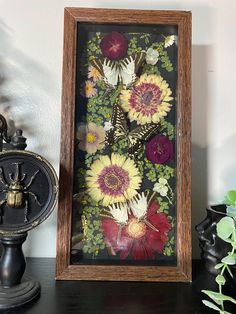 there is a framed painting with flowers in it on the shelf next to a clock