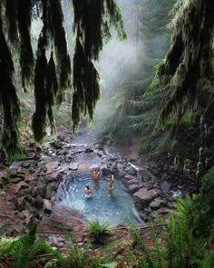 three people sitting in a hot tub surrounded by rocks and trees, with the water running through them