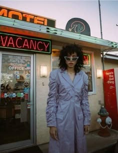 a woman standing in front of a motel wearing sunglasses and a purple trench coat with the word vacancy written on it