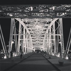 black and white photograph of people walking across a bridge at night with city lights in the background