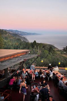 people are sitting at tables on top of a hill overlooking the water and hills in the distance