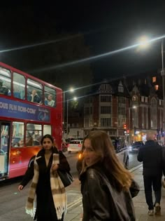 two women walking down the street in front of a red double decker bus at night