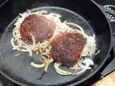 two meat patties being cooked in a frying pan on the stove with onions