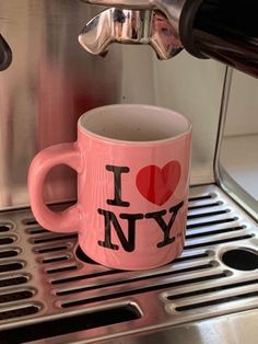 a pink coffee mug with i love new york on it sitting in front of a silver espresso machine