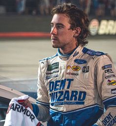 a man with long hair standing in front of a car holding a white jersey and looking off into the distance