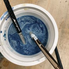 two paintbrushes in a white bowl with blue liquid and water on the floor